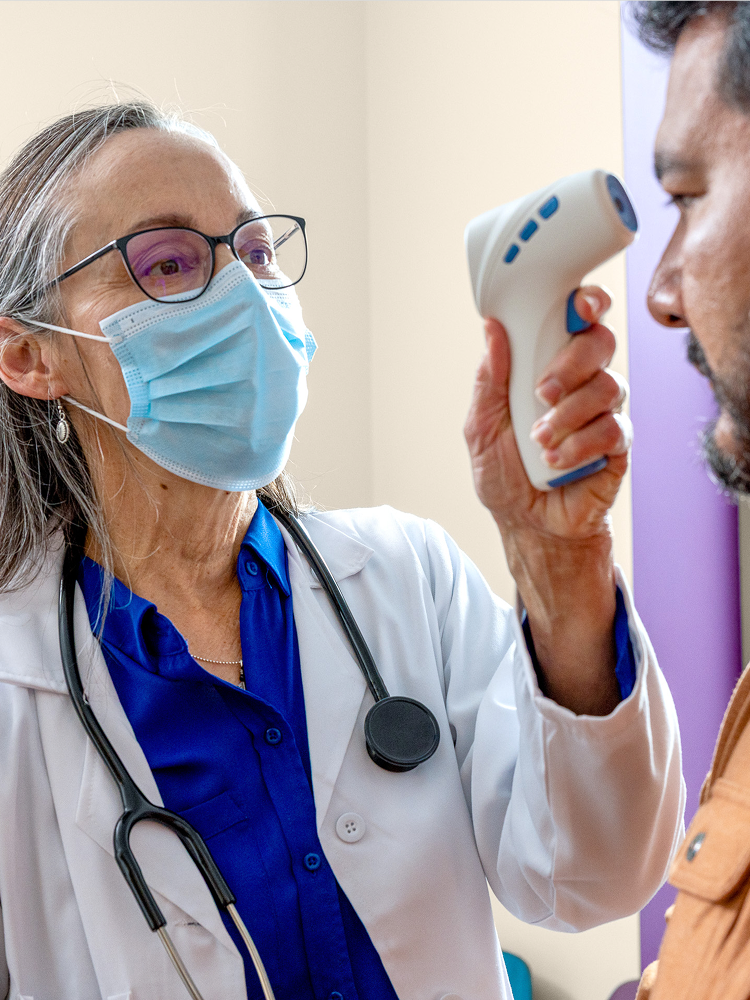 Masked clinician in a white coat checking a patient's temperature with a no-touch forehead thermometer