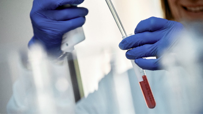 Gloved technician transferring a sample into a test tube with a pipette in a lab
