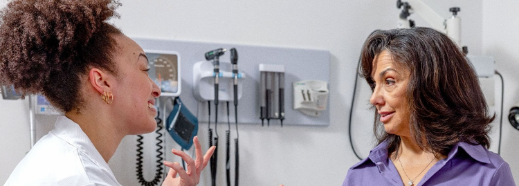 Clinician in a white coat gesturing while talking with a seated patient in an exam room