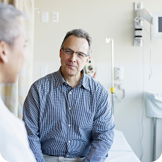 Man in a checkered shirt sitting in a hospital room while talking with another person in the foreground