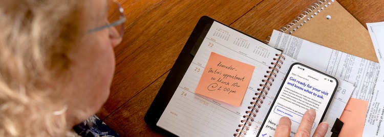 Woman reviewing a doctor's appointment reminder on a sticky note in a planner while on a PfizerForAll Prepare for Care page on her phone