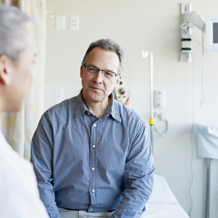 Man in a checkered shirt sitting in a hospital room while talking with another person in the foreground