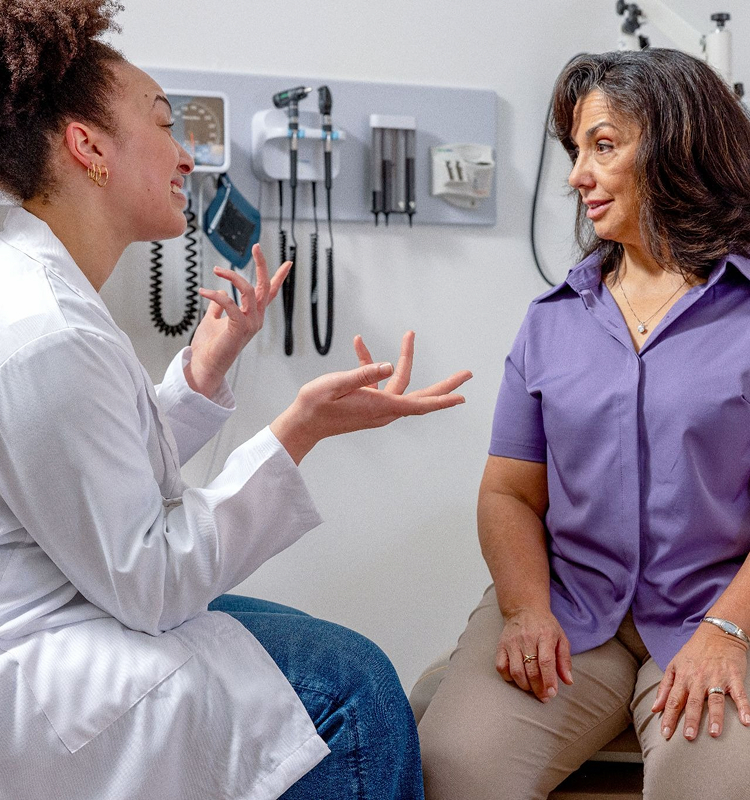 Clinician in a white coat gesturing while talking with a seated patient in an exam room
