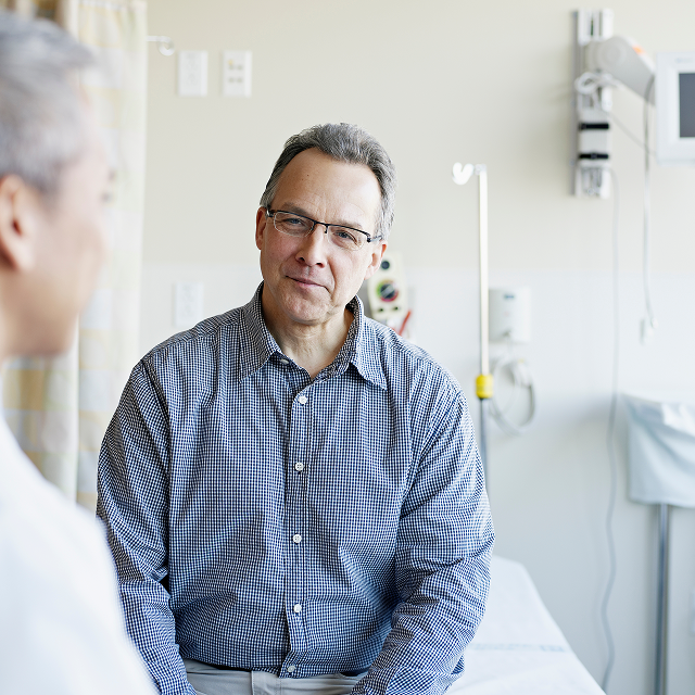 Man in a checkered shirt sitting in a hospital room while talking with another person in the foreground