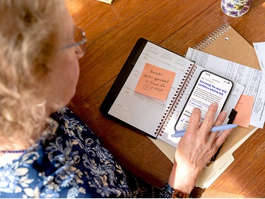 Woman reviewing a doctor's appointment reminder on a sticky note in a planner while on a PfizerForAll Prepare for Care page on her phone