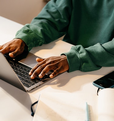 Person in a green sweatshirt typing on a laptop at a desk with a phone nearby