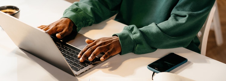Person in a green sweatshirt typing on a laptop at a desk with a phone nearby